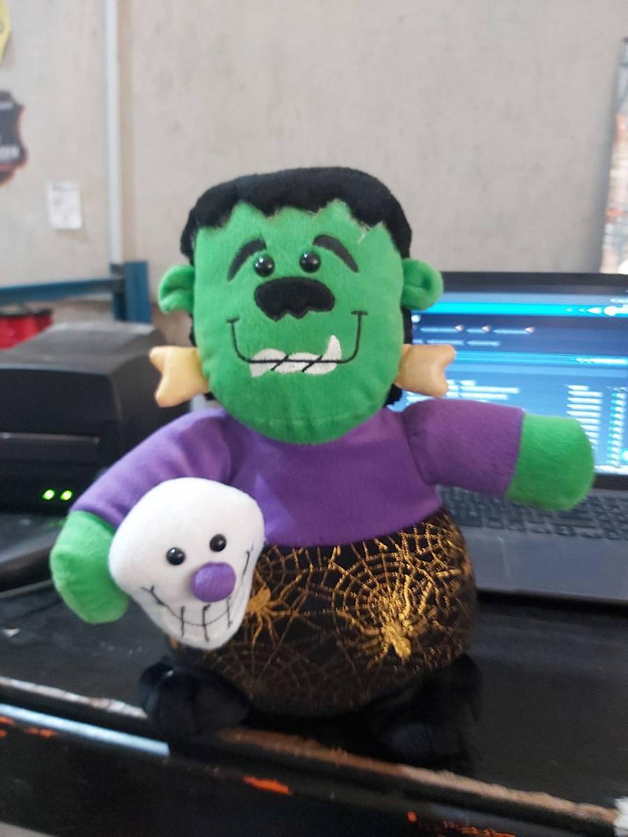 Boneco de pelúcia verde com roupa roxa e detalhe de caveira branca